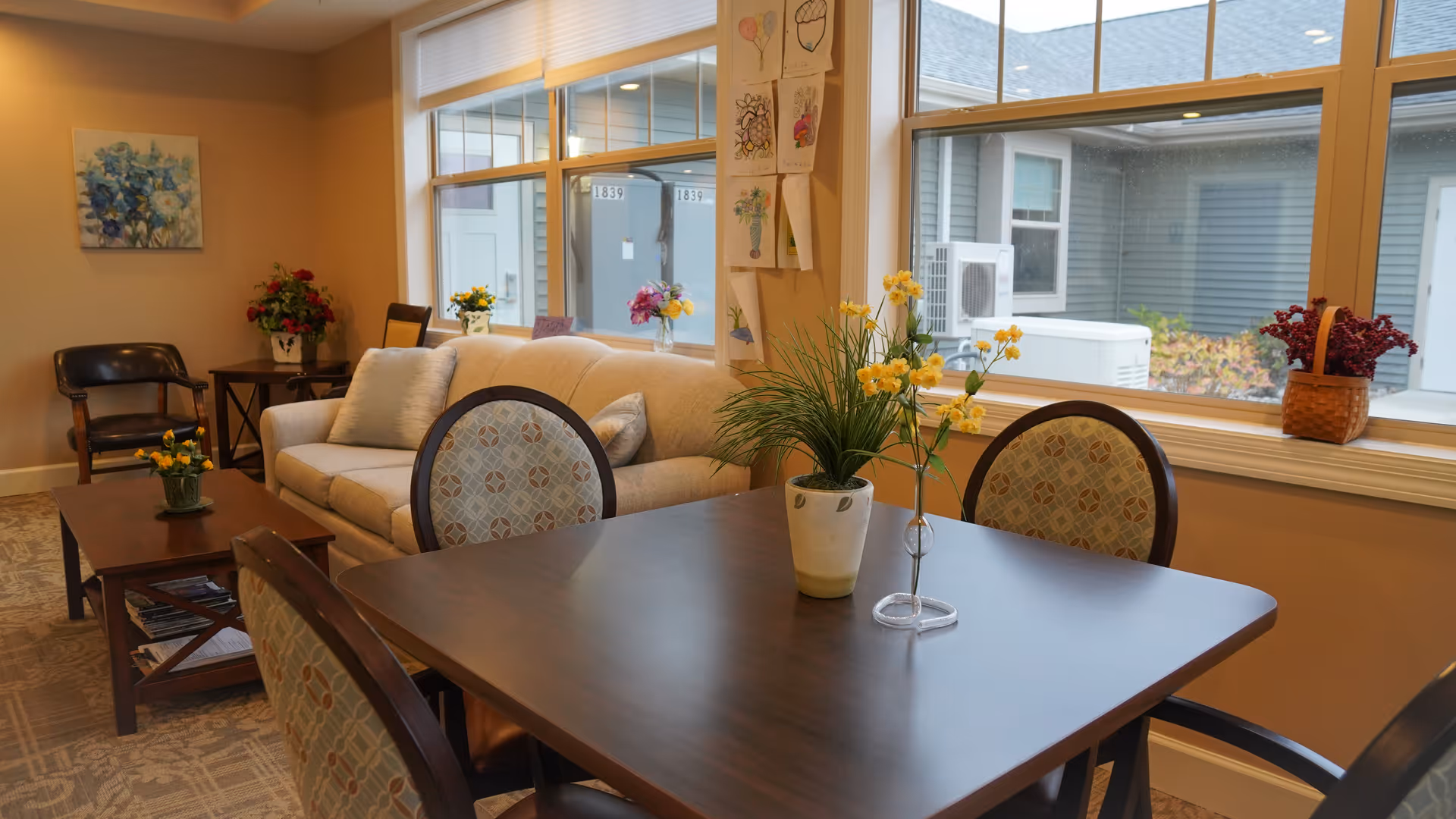 Bright communal living room with a wooden dining table and chairs, a sofa and coffee table, and floral decorations by large windows.