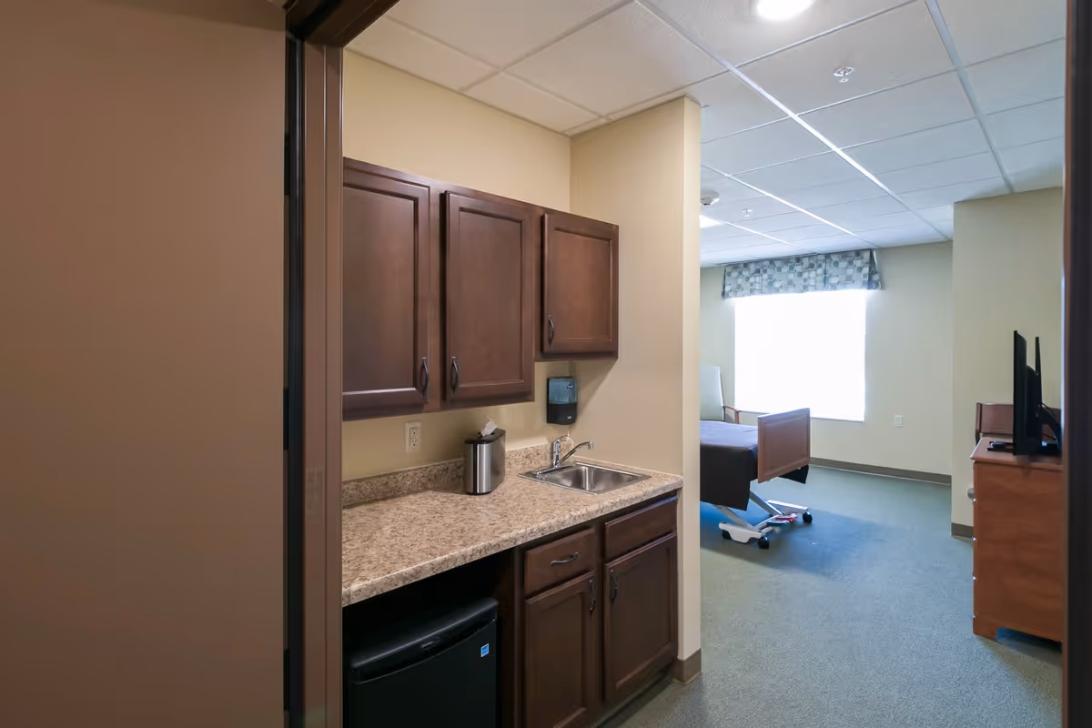 Interior view of a senior living facility room at Gracepointe Crossing Gables showing a small kitchenette area with dark wood cabinets, a countertop with a sink, and a mini refrigerator. In the background, there is a hospital-style bed near a large window with a patterned valance, and a wooden dresser with a TV on top.