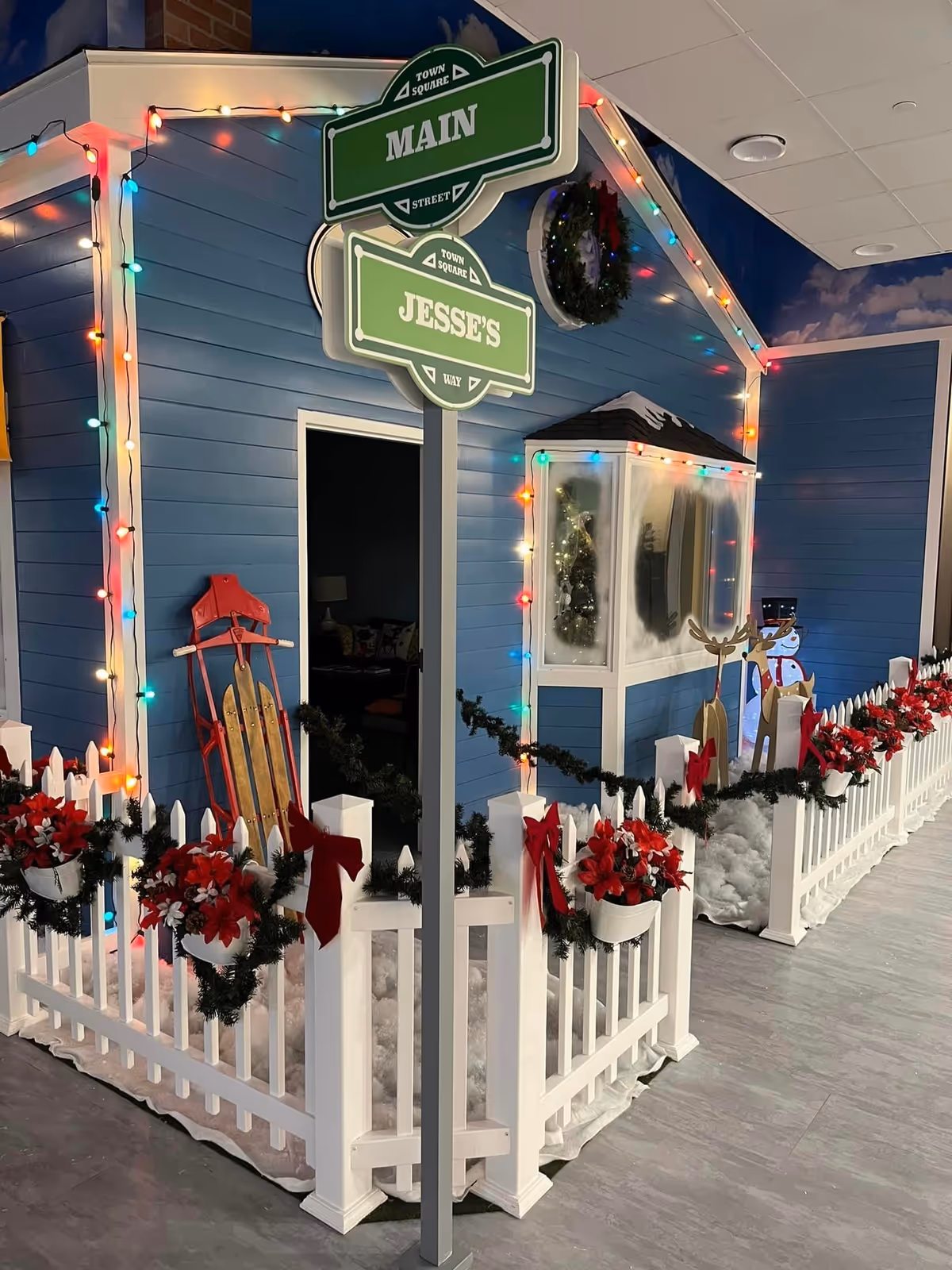 Indoor holiday-themed display featuring a small blue house decorated with multicolored string lights, a white picket fence adorned with red bows and poinsettias, a red sled leaning against the house, and festive decorations including a wreath, reindeer cutouts, and a snowman figure.