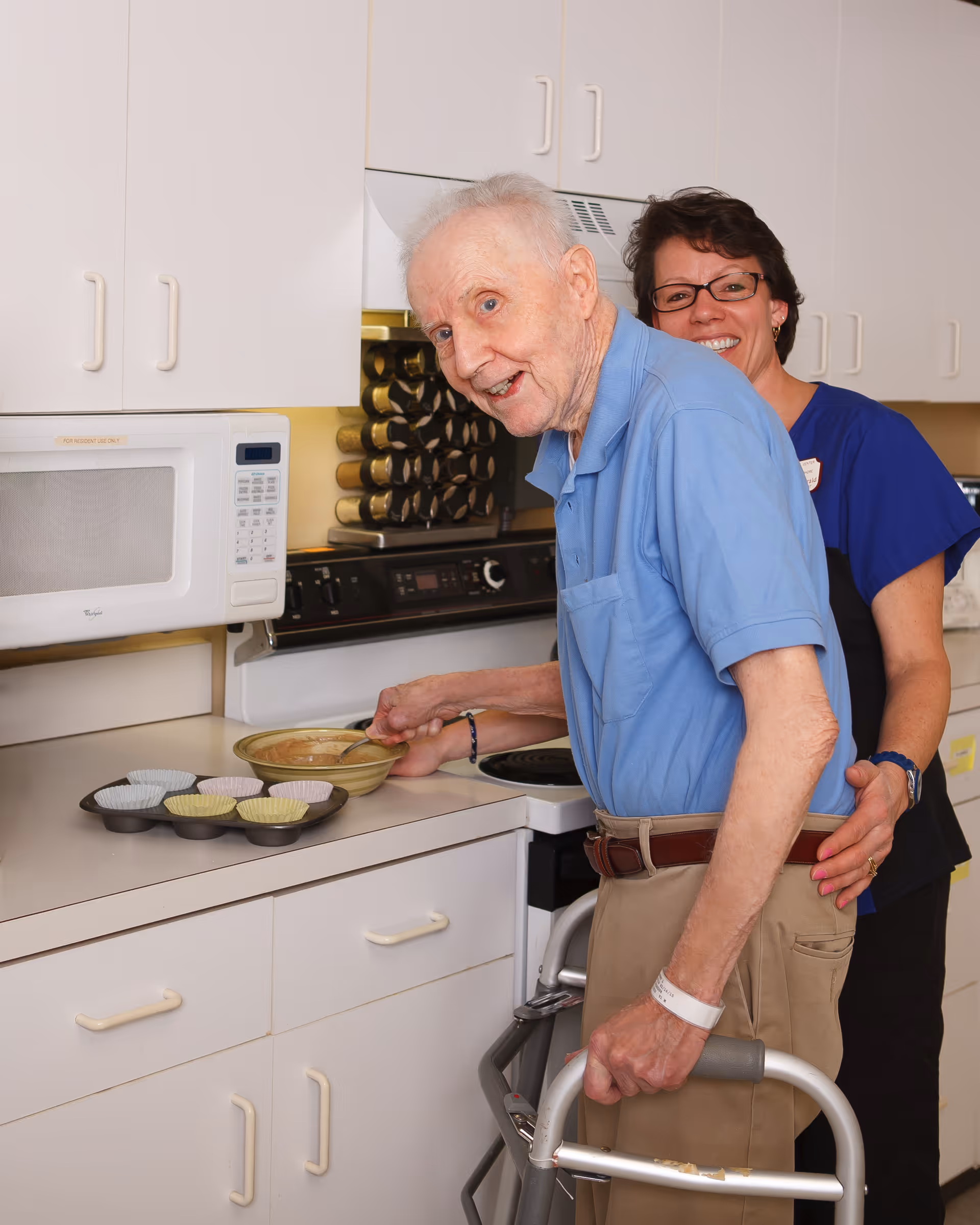An elderly man using a walker is mixing batter in a bowl on a kitchen counter, with a woman standing behind him smiling and offering support. The kitchen has white cabinets, a microwave, and a stove with a spice rack in the background.
