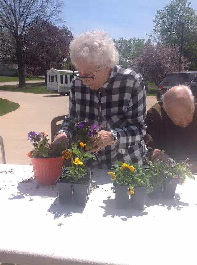 An elderly woman wearing glasses and a black-and-white checkered shirt is planting flowers in pots on a white table outdoors. Another elderly person is seated next to her, also engaged in the activity. Trees, a vehicle, and a paved path are visible in the background under a clear sky.