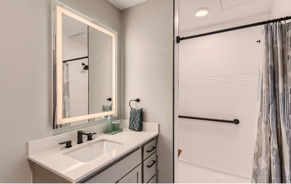 Modern bathroom with a white countertop vanity featuring a rectangular sink and black faucet fixtures. Above the vanity is a large rectangular mirror with built-in lighting. To the right, there is a shower area with a white wall, black grab bars, and a patterned shower curtain.