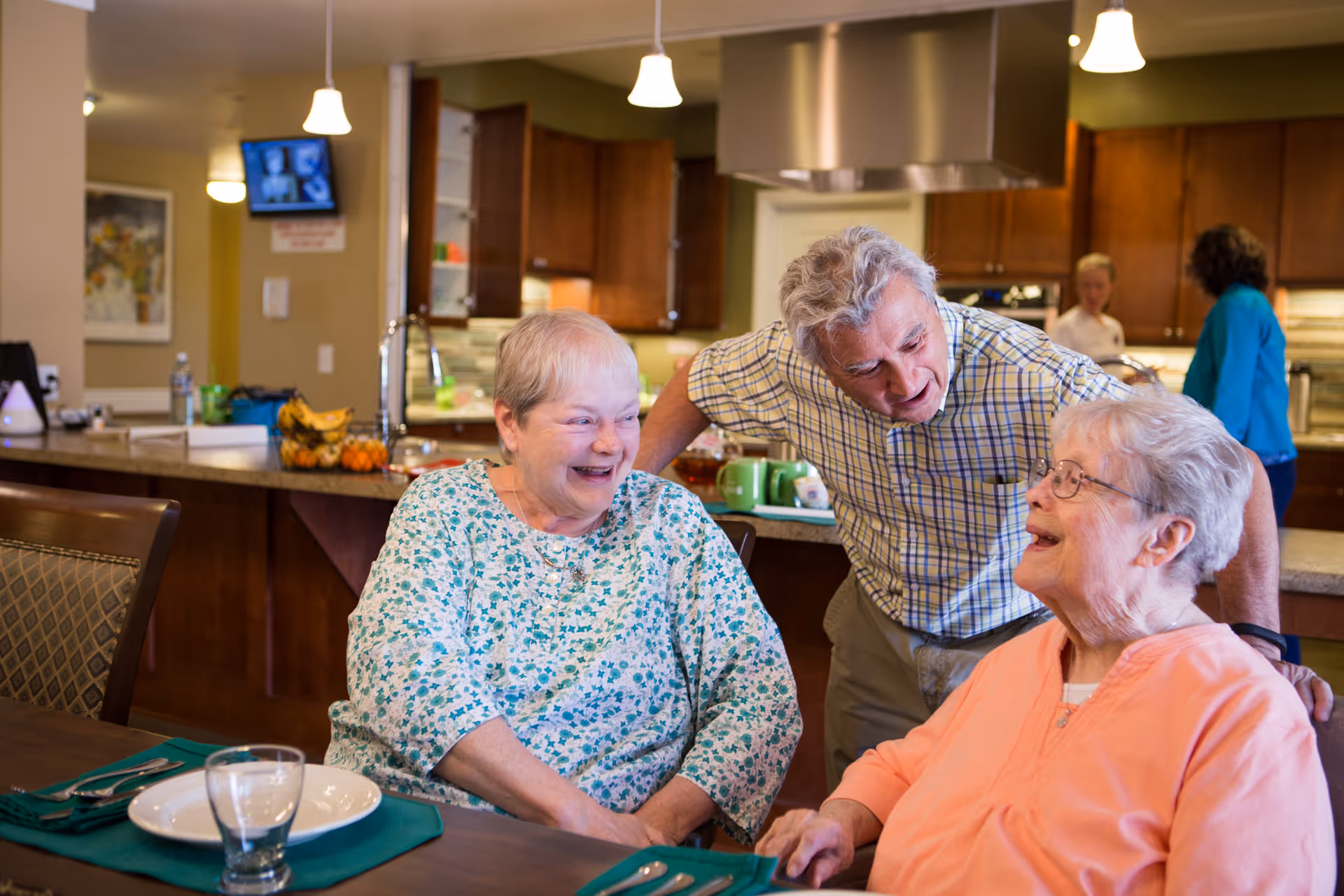 Three elderly residents laughing and talking around a dining table in a communal kitchen/dining area.