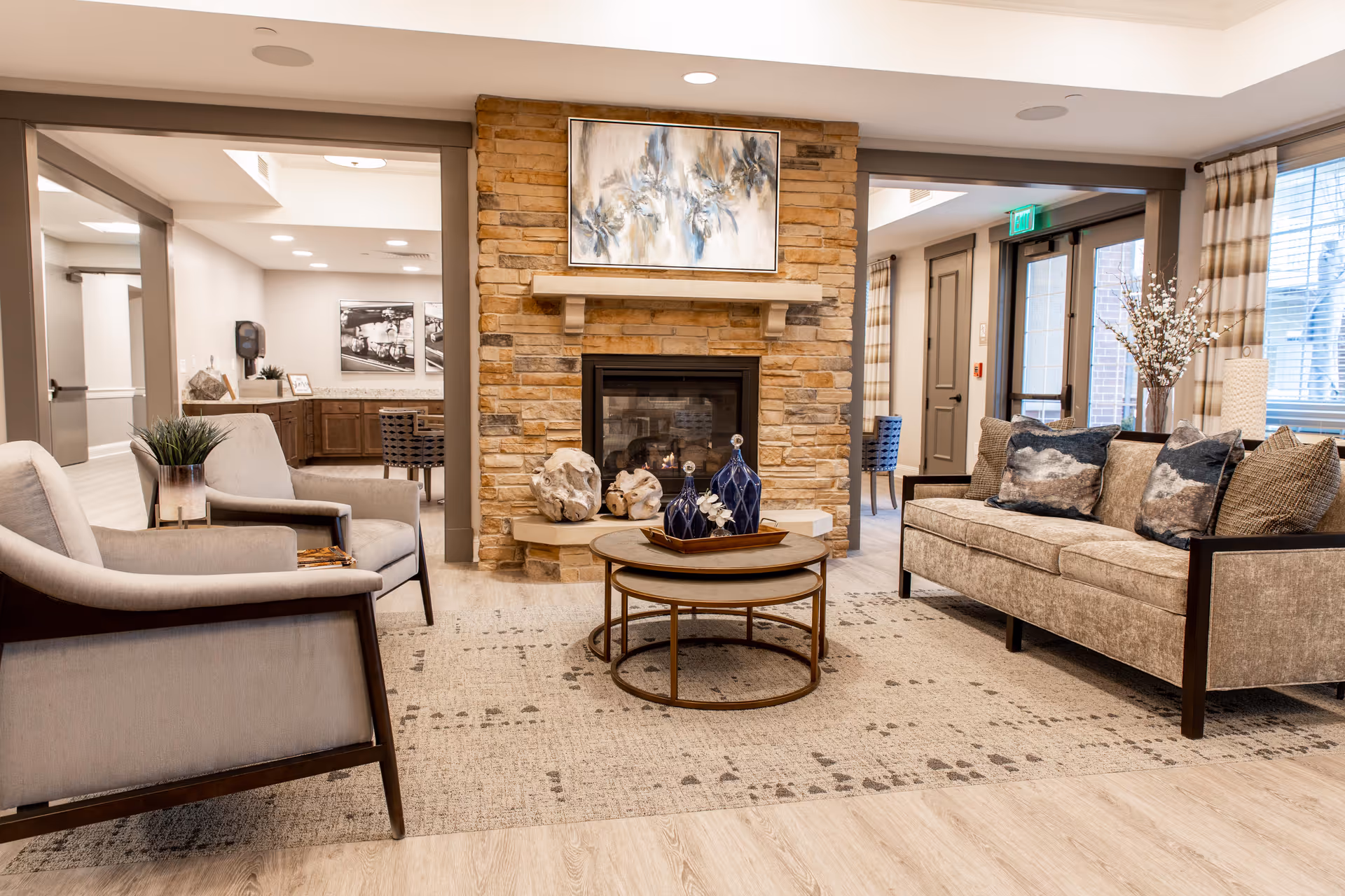 A cozy senior living facility lounge area featuring a stone fireplace with a painting above it, two beige armchairs, a beige sofa with decorative pillows, a round coffee table with blue vases, and large windows with curtains allowing natural light to fill the room.