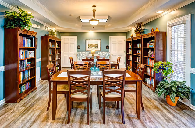Well-lit communal dining and reading room with wooden tables and chairs, bookshelves, and plants.