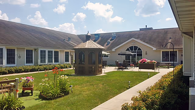 Outdoor courtyard area of a senior living facility with a wooden gazebo in the center, surrounded by green grass, flower beds, and a paved walkway. The building has large windows and a sloped roof under a partly cloudy sky.