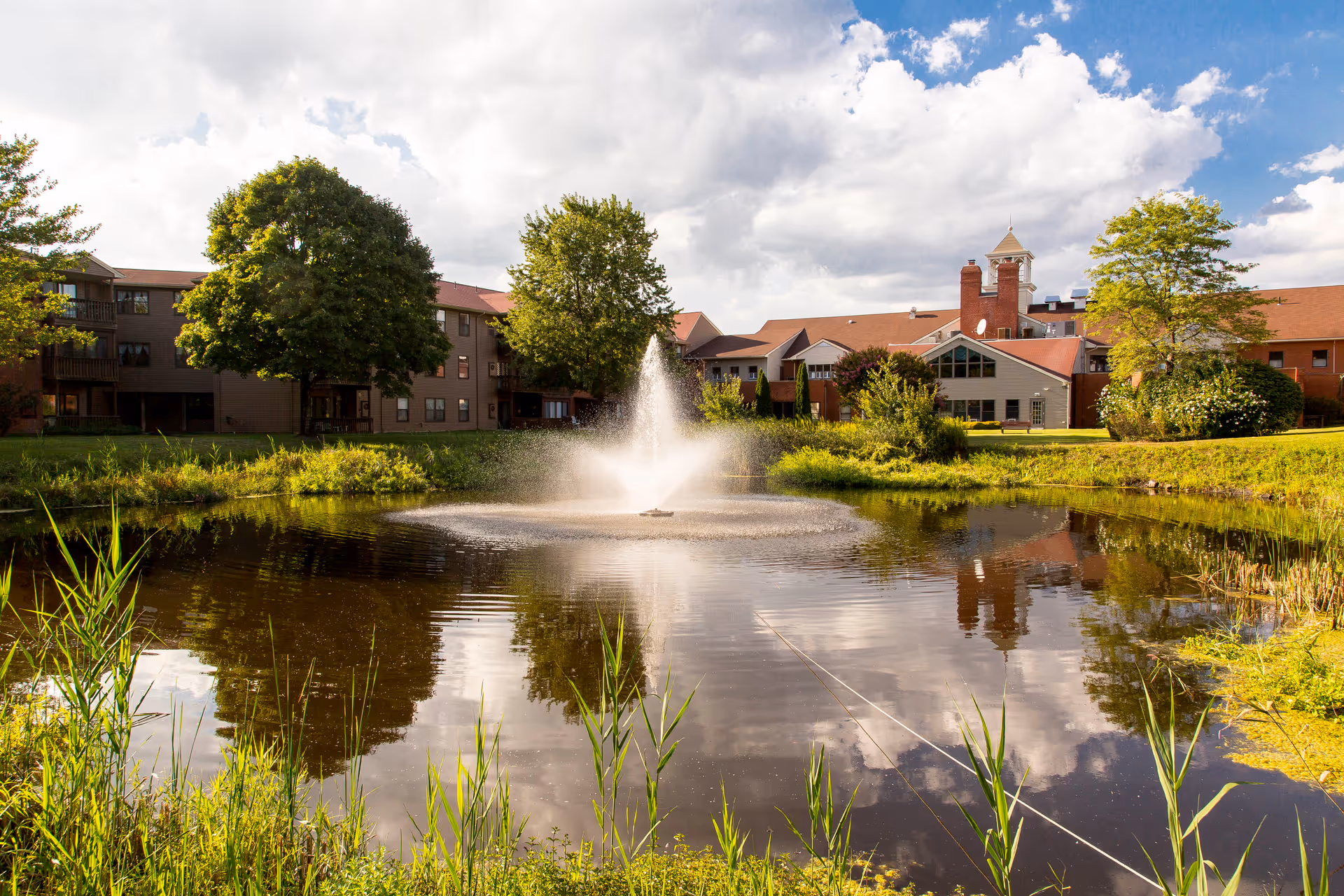 A scenic view of a pond with a water fountain in the center, surrounded by green grass and trees. In the background, there is a large building with a red roof and multiple windows under a partly cloudy sky.