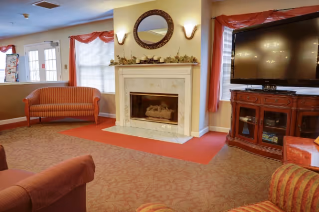 Common area living room with a fireplace and mantel, round mirror above it, a large TV on a wooden cabinet, and upholstered sofas.