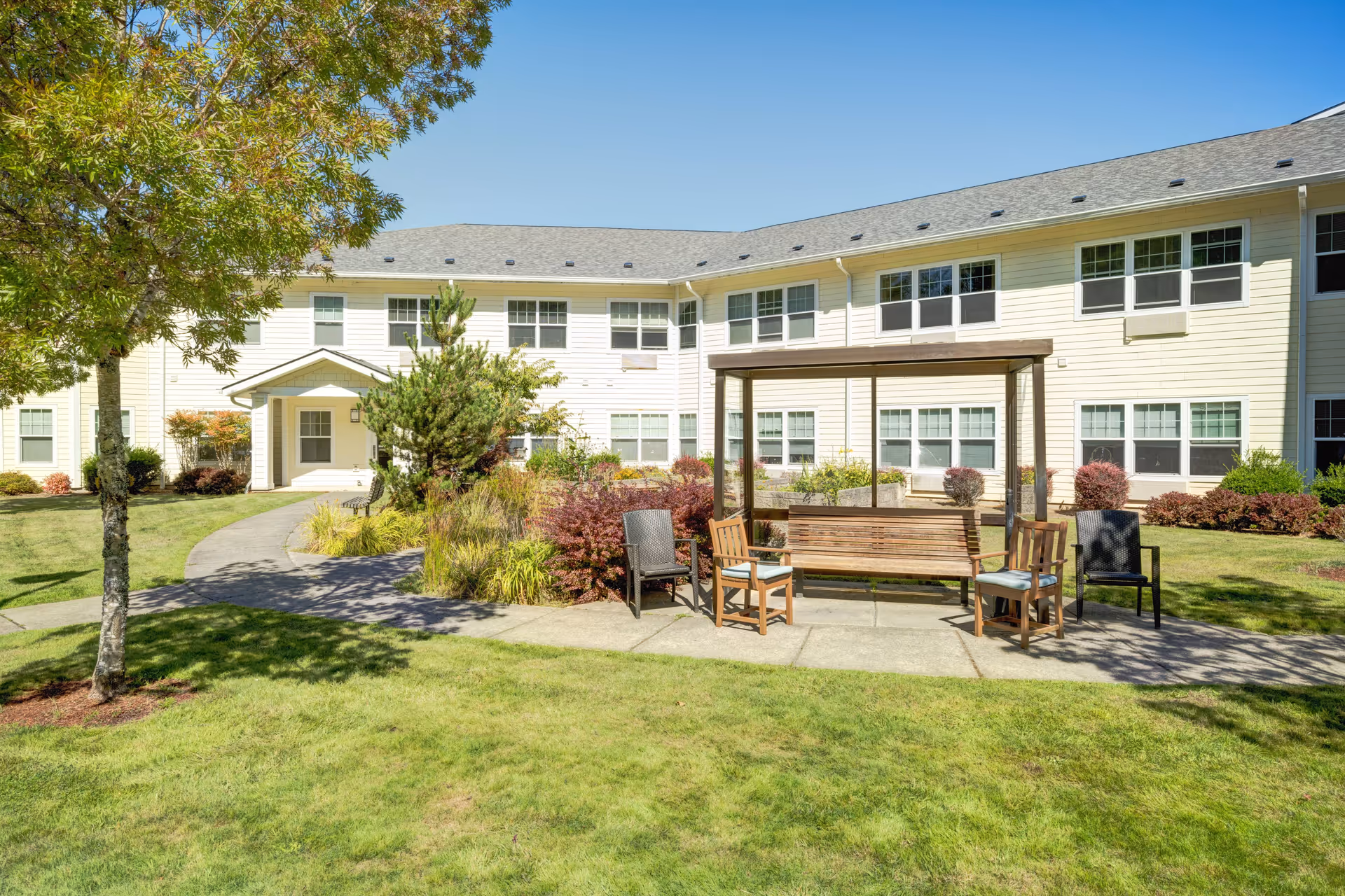 Outdoor seating area with a wooden swing bench and several chairs arranged on a concrete patio surrounded by grass and landscaping. In the background, there is a two-story light yellow building with multiple windows under a clear blue sky.