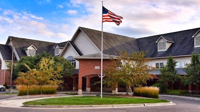 Exterior view of a senior living facility building with a large American flag on a flagpole in front. The building has a brick and white facade with multiple gabled roofs and dormer windows. There are trees and landscaped bushes around the entrance area under a partly cloudy sky.