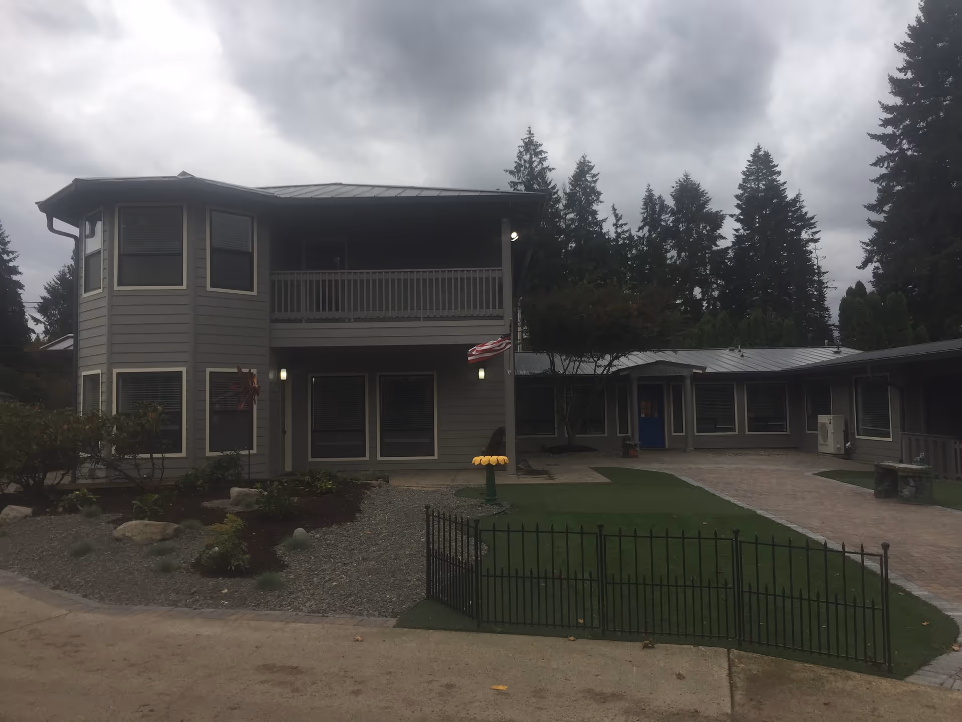 Exterior view of a two-story gray building with multiple windows and a balcony on the upper floor. The building is surrounded by a landscaped area with shrubs, rocks, and a small fenced-in lawn. There is a paved walkway leading to a blue door under a small porch. Tall evergreen trees are visible in the background under a cloudy sky.