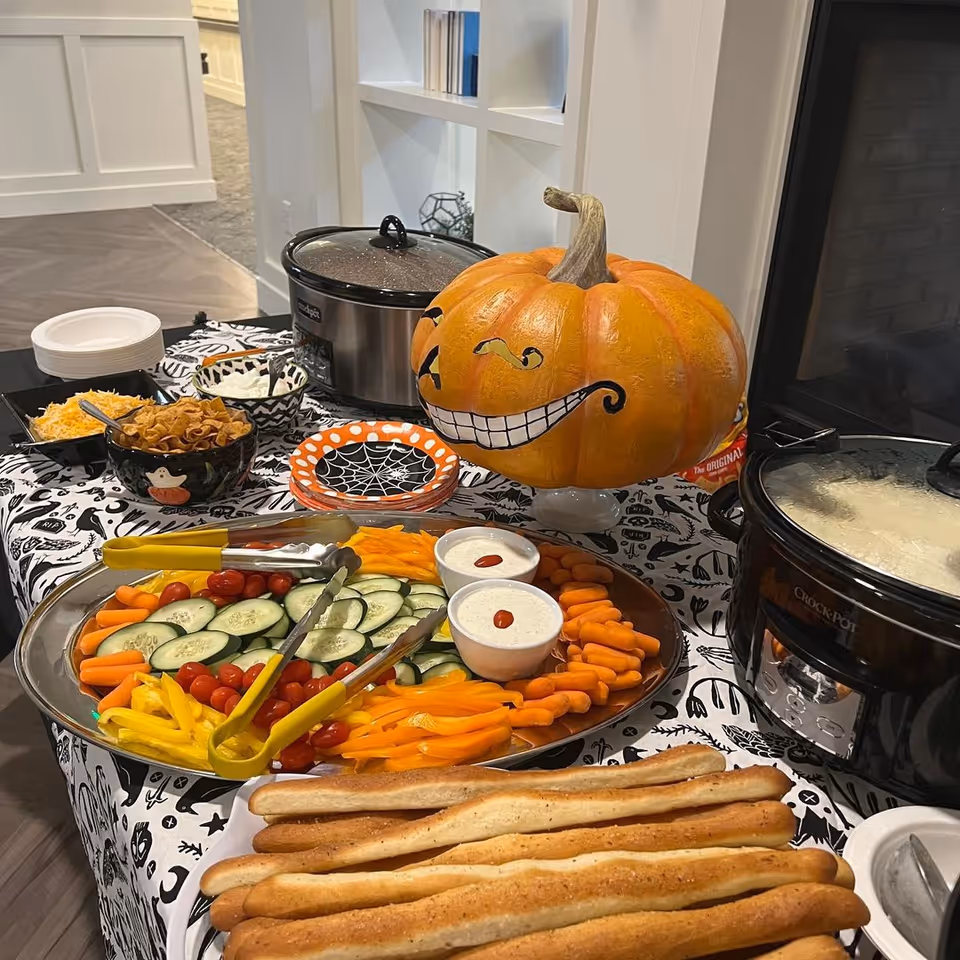 A festive food buffet table with a Halloween theme, featuring a large pumpkin painted with a mischievous smiling face, a platter of fresh vegetables including cucumbers, cherry tomatoes, baby carrots, and yellow bell peppers with two bowls of ranch dressing, breadsticks, shredded cheese, and other toppings. Two crockpots are also visible on the table, which is covered with a black and white Halloween-themed tablecloth.