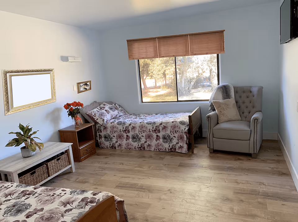 A bright and clean bedroom in an assisted living facility featuring two single beds with floral bedspreads, a wooden nightstand with a vase of red flowers, a cushioned armchair with a pillow and throw blanket, a large window with wooden blinds, a wall-mounted TV, a decorative mirror, and a small table with two wicker baskets underneath.
