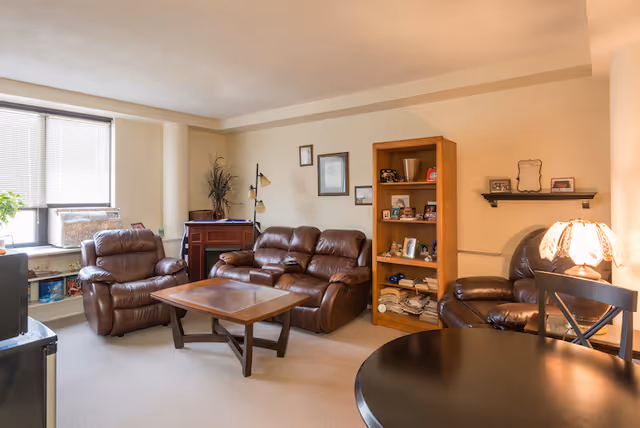 A cozy living room with beige walls and carpet, featuring a brown leather sofa, two matching armchairs, a wooden coffee table, a wooden bookshelf filled with books and framed photos, a floor lamp, and a round black table with chairs. A window with blinds allows natural light into the room.