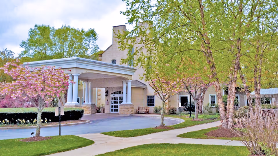 Entrance of Saint Elizabeth Home Skilled Nursing & Rehabilitation Center with a covered driveway, surrounded by green trees and blooming pink flowers, and a sidewalk leading to the entrance.