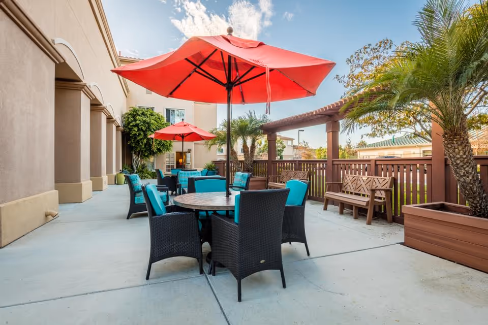 Outdoor patio area with round tables and black wicker chairs featuring turquoise cushions. Each table has a large red umbrella providing shade. The patio is adjacent to a beige building with arched columns. There are palm trees and wooden benches along a brown fence, under a partly cloudy sky.