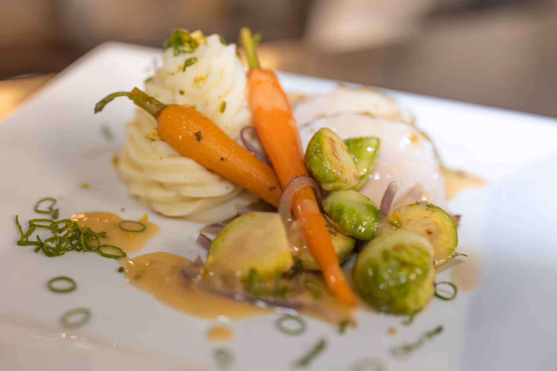 A plated gourmet meal featuring two baby carrots, mashed potatoes garnished with herbs, halved Brussels sprouts, and a piece of white fish with sauce on a white square plate.