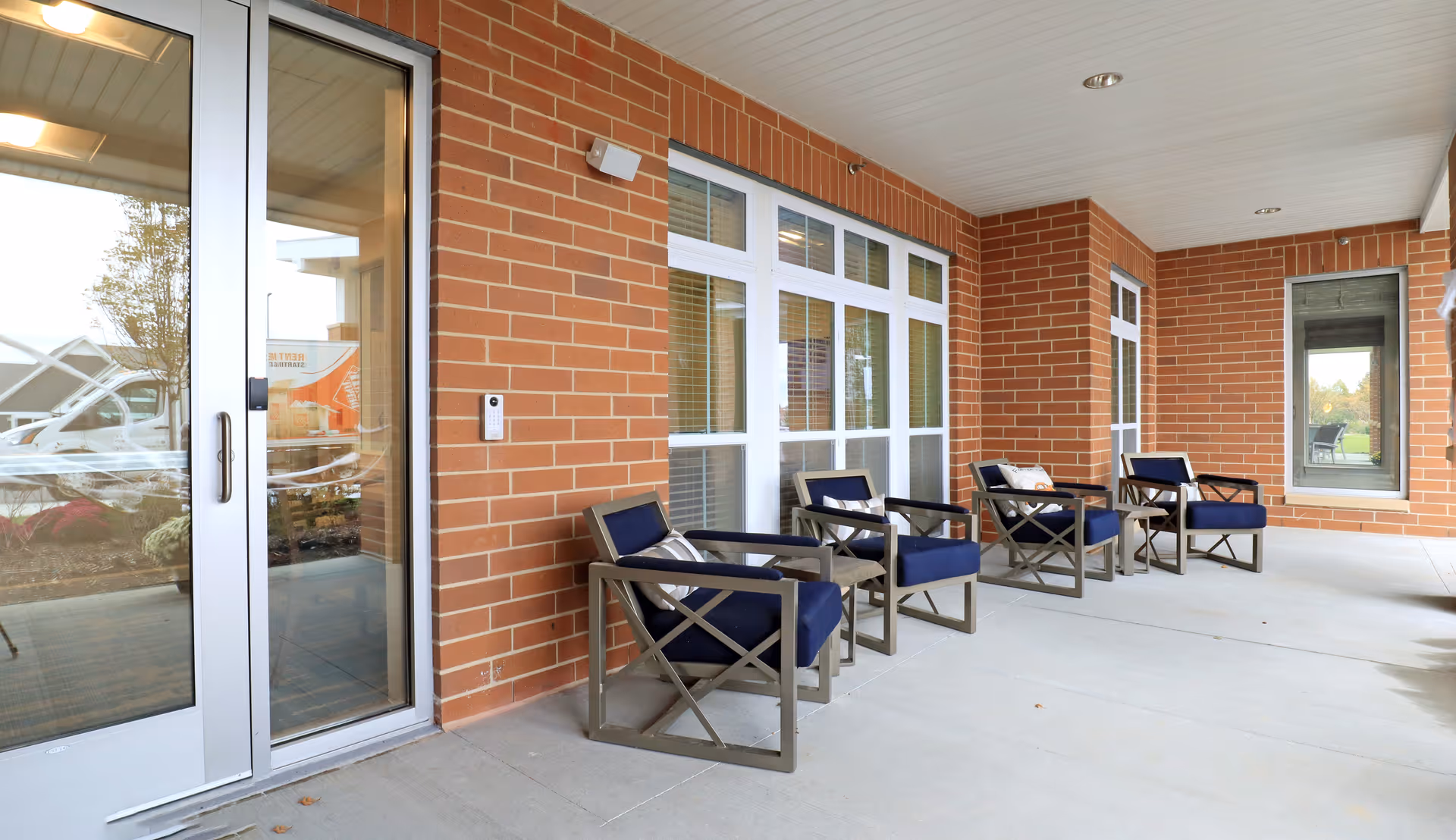 Covered brick exterior patio with a row of navy cushioned chairs beside large windows and glass entry doors.