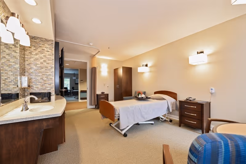 A well-lit senior living facility bedroom with a single bed covered in a light gray blanket. The room features a wooden wardrobe, a bedside table with a telephone, and a small table with a tray of flowers and a cup. There is a sink with a countertop and a large mirror on the left side. The walls are painted beige, and the floor is carpeted. A chair with blue upholstery is partially visible in the foreground.