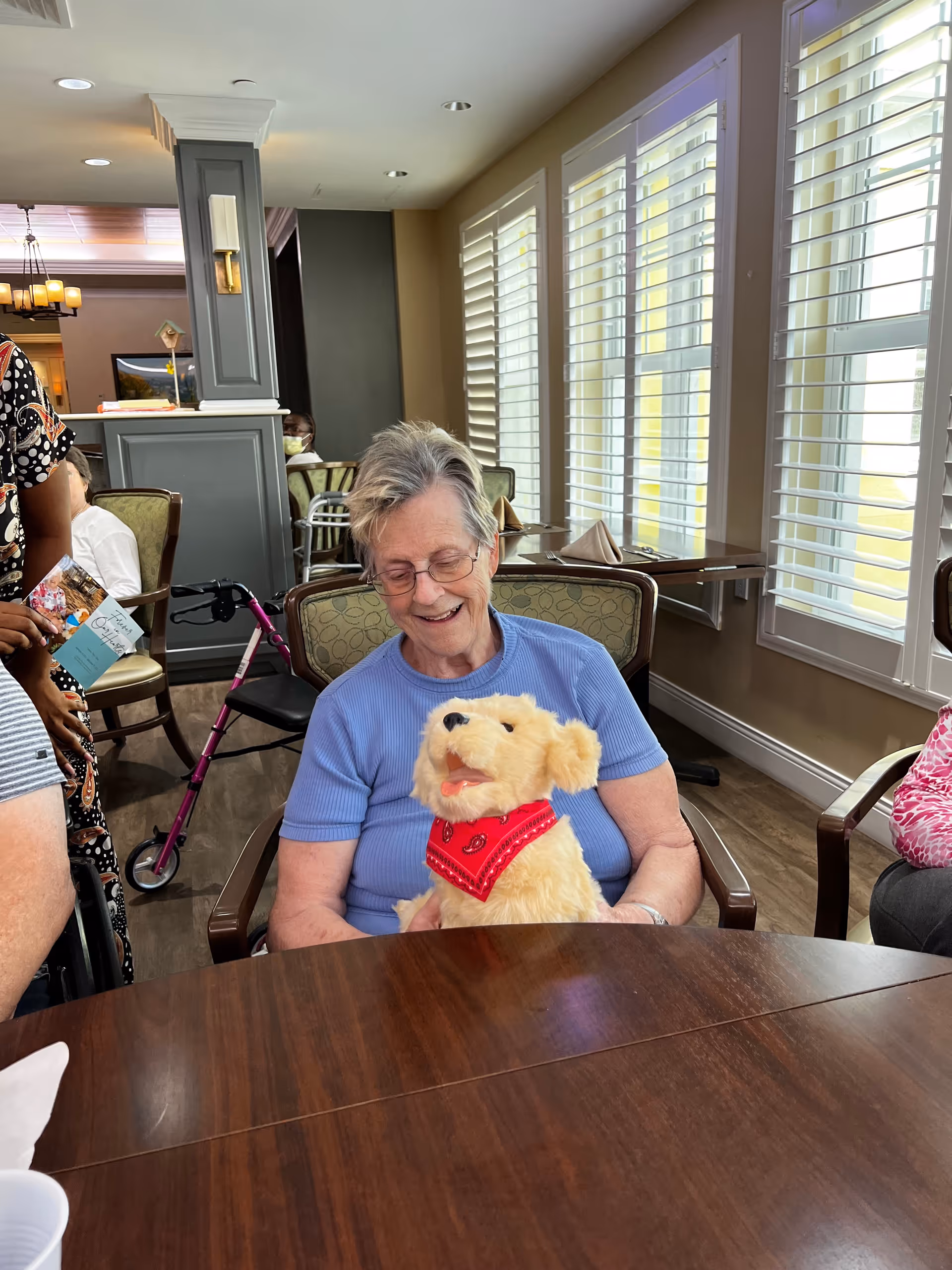 An elderly woman wearing glasses and a blue shirt is sitting at a wooden table in a well-lit room with large windows and white shutters. She is smiling and holding a plush dog toy wearing a red bandana. Other people and chairs are visible in the background, along with a pink walker.