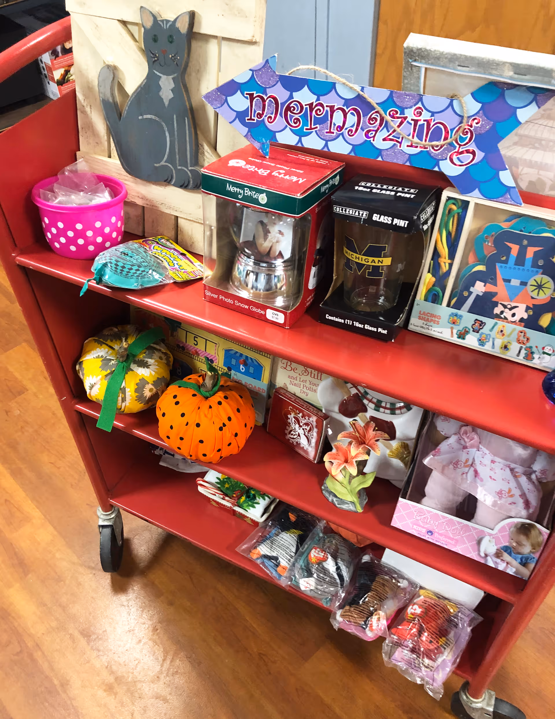 A red rolling cart filled with various decorative items including a wooden cat figure, a 'mermazing' sign shaped like a fish, a silver photo snow globe, a Michigan glass pint, small pumpkins, a flower figurine, and packaged toys and socks.