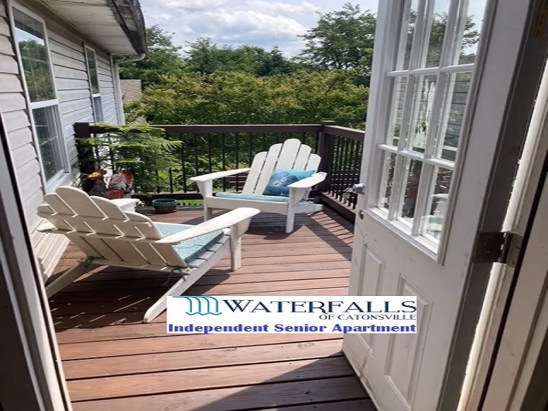 View through an open door onto a wood deck balcony with two white Adirondack chairs, cushions, and potted plants overlooking trees.