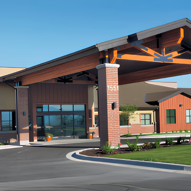 Covered entrance canopy and driveway in front of a single-story senior living facility with brick pillars and landscaped lawn.