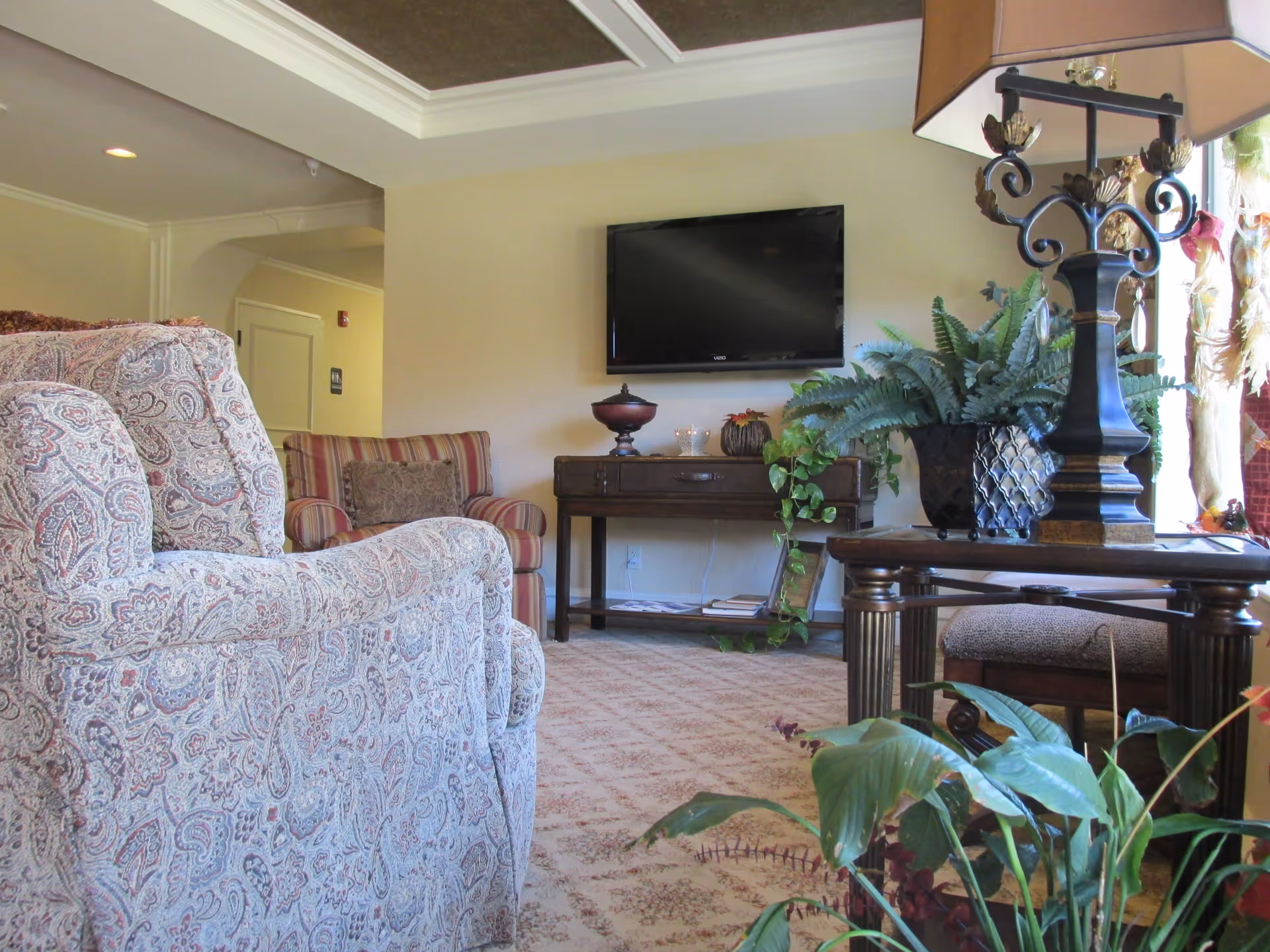 A cozy living room area with patterned upholstered armchairs, a wooden table with a decorative lamp and potted plants, a flat-screen TV mounted on a beige wall, and a console table with decorative items and more plants underneath. The room has a carpeted floor and a tray ceiling with recessed lighting.