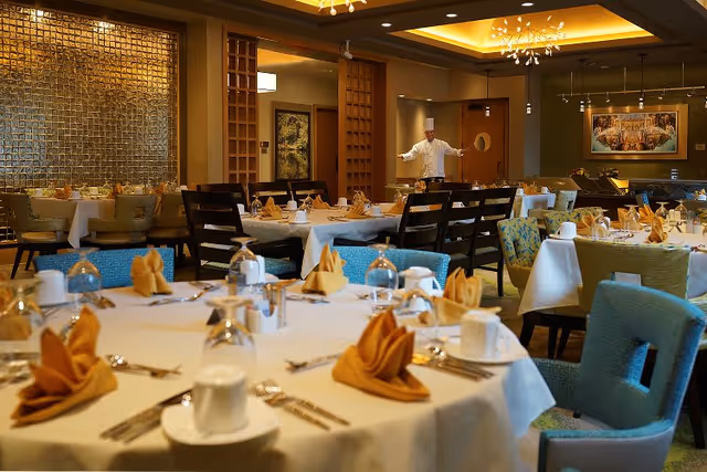 A dining room in a senior living facility with tables set with white tablecloths, folded orange napkins, cups, glasses, and silverware. Chairs in various colors surround the tables. A chef in a white uniform and hat stands in the background near the entrance, welcoming guests. The room has warm lighting and decorative elements on the walls.