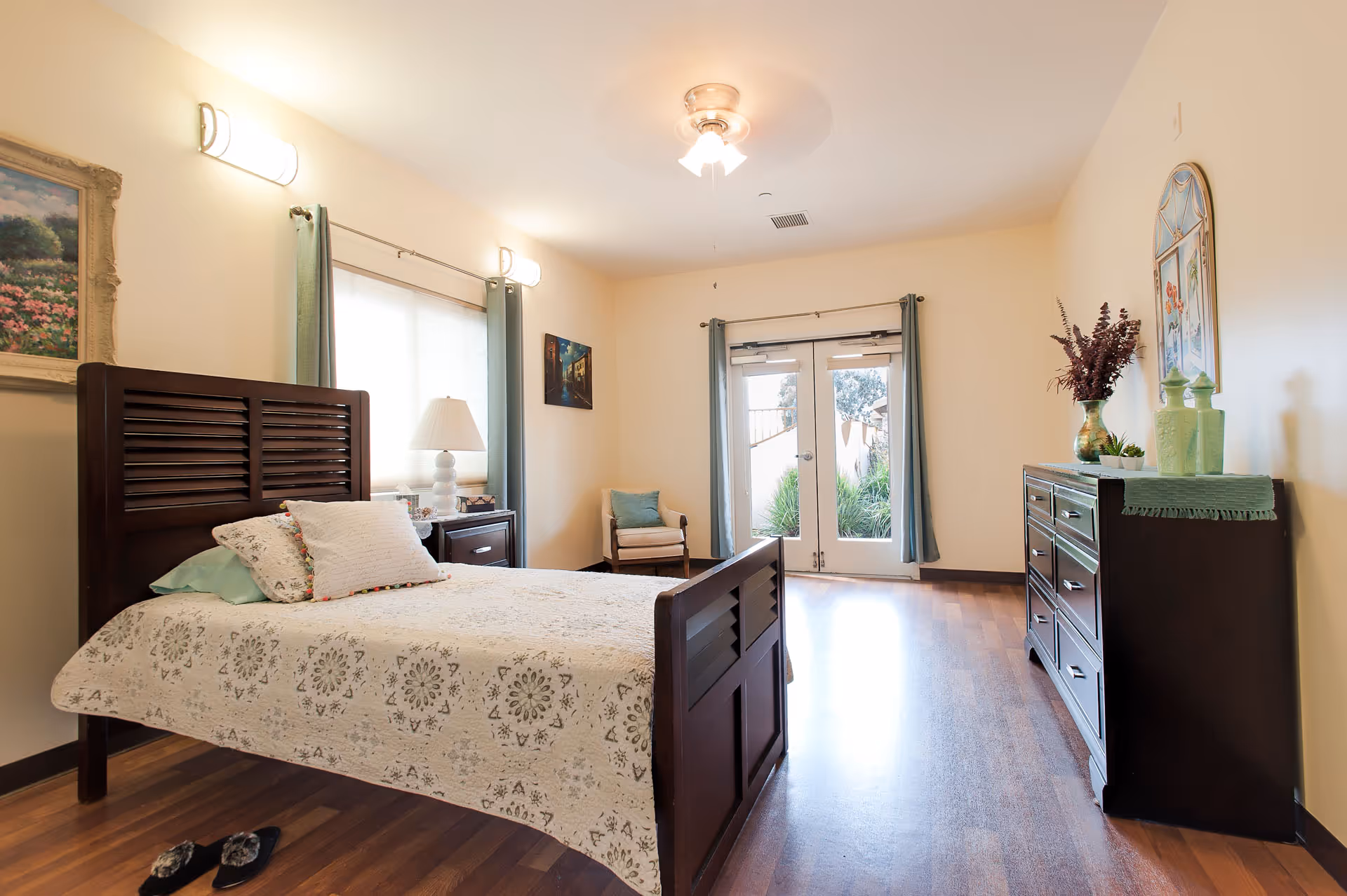 A bright and cozy bedroom in a senior assisted living facility featuring a wooden bed with patterned bedding, a nightstand with a lamp, a chair in the corner, a dresser with decorative items, and double glass doors leading outside. The room has light-colored walls, wooden flooring, and curtains on the windows and doors.