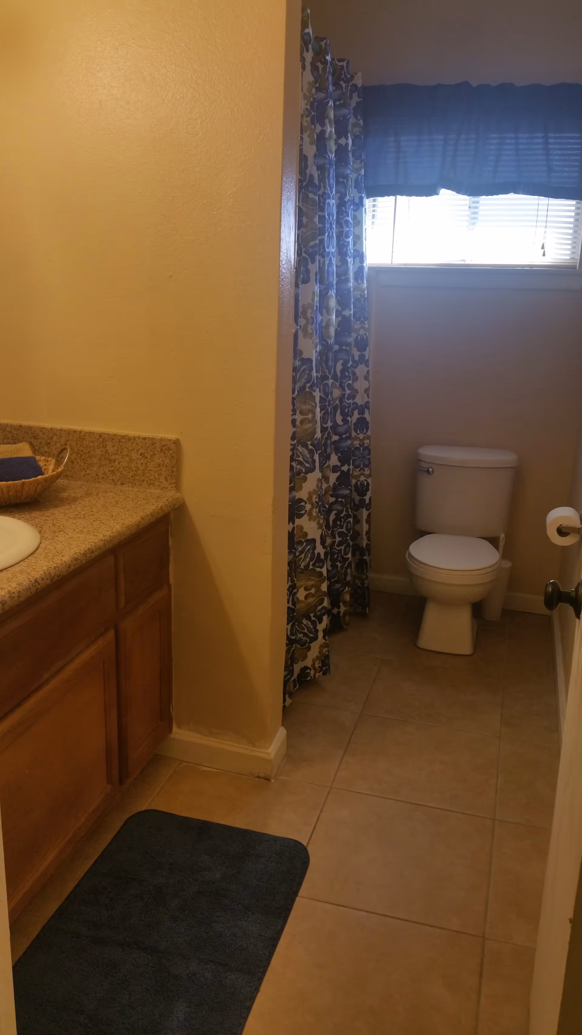 A small bathroom with beige tiled floor, a white toilet, a wooden cabinet with a granite countertop and sink, a blue bath mat, and a window with blue and white floral curtains.