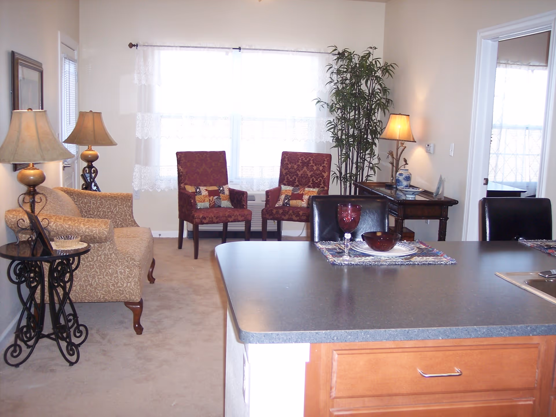 A cozy living room area with two patterned armchairs and a beige sofa, illuminated by two table lamps. A small side table with decorative iron legs is next to the sofa. In the foreground, there is a kitchen island with a place setting including a red wine glass, bowl, and plate. A tall green plant and a wooden console table with a lamp and decorative items are against the wall near a window with sheer white curtains.