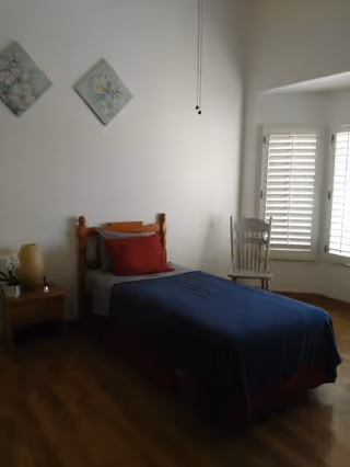 A simple bedroom with a single wooden bed covered with a blue blanket and red pillows. Next to the bed is a small wooden nightstand with a lamp and a small plant. There are two floral paintings hanging on the white wall above the bed. A wooden chair is placed near a window with white plantation shutters, allowing natural light into the room. The floor is wooden.