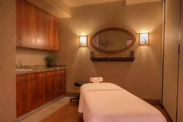 A serene massage therapy room with a massage table covered in a white sheet, a small black stool, wooden cabinets with granite countertops on the left, an oval mirror on the wall, and two wall-mounted lights providing warm lighting.