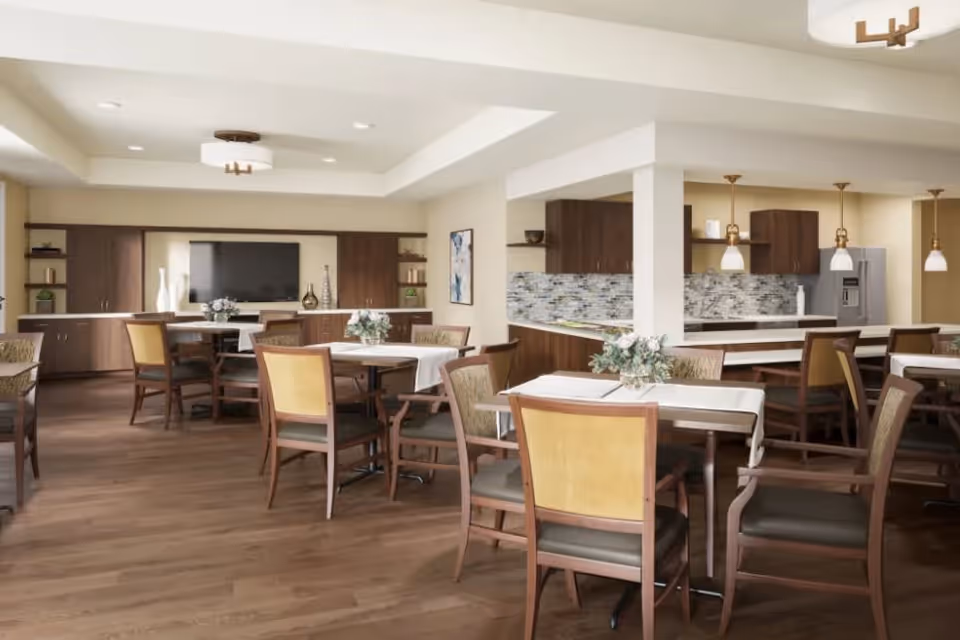 A spacious dining area with multiple tables and chairs arranged neatly. The room features wooden flooring, a modern kitchen area with pendant lights, and a large flat-screen TV mounted on the wall. The tables are decorated with small floral centerpieces.