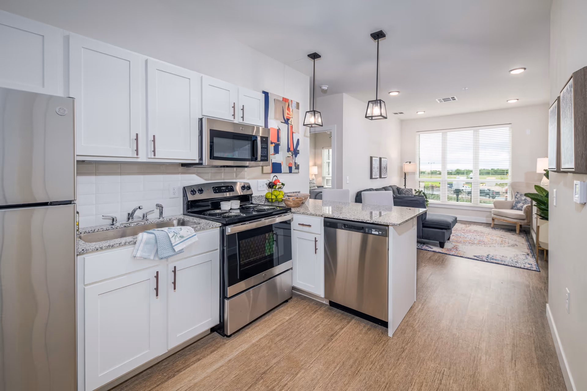 Modern kitchen with white cabinets, stainless steel refrigerator, oven, microwave, and dishwasher. The kitchen island has a granite countertop with two chairs. In the background, there is a living room area with a large window, a gray sectional sofa, an armchair, and a colorful rug.