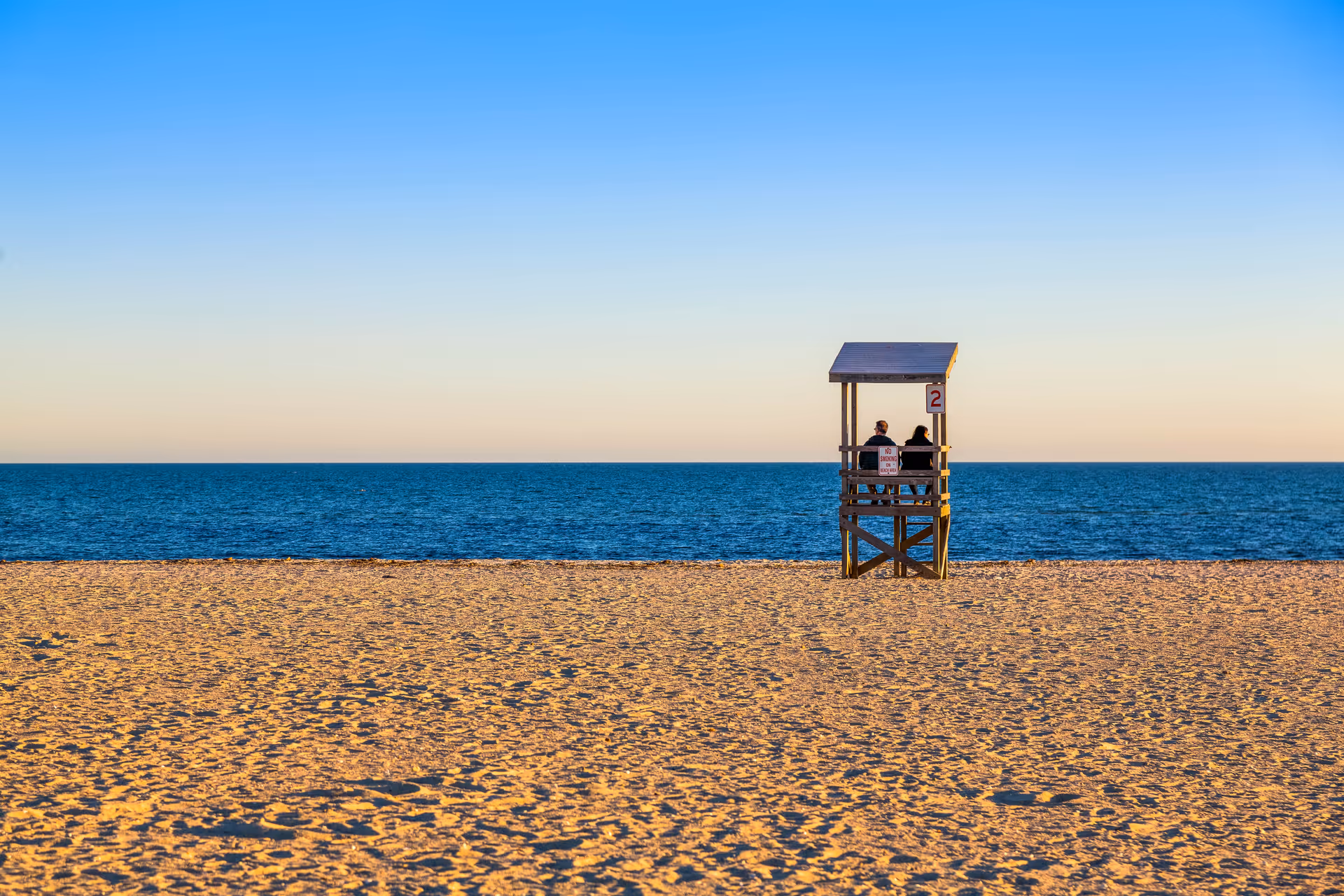 A lifeguard stand with two people sits on a wide sandy beach facing a calm ocean under a clear sky.