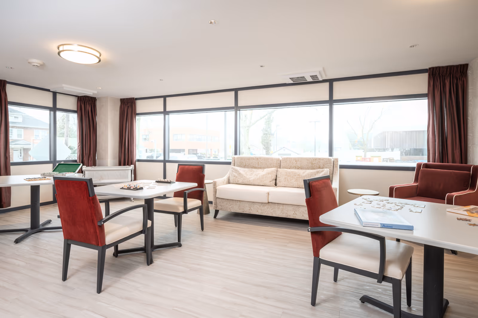A bright and spacious common area in a senior living facility with large windows letting in natural light. The room features a beige sofa, red cushioned chairs, and tables with board games and puzzles on them. The floor is light wood, and there are dark red curtains on the windows.