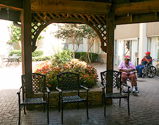 Covered outdoor courtyard with decorative metal chairs around a central planter and two residents near the building, one in a wheelchair.