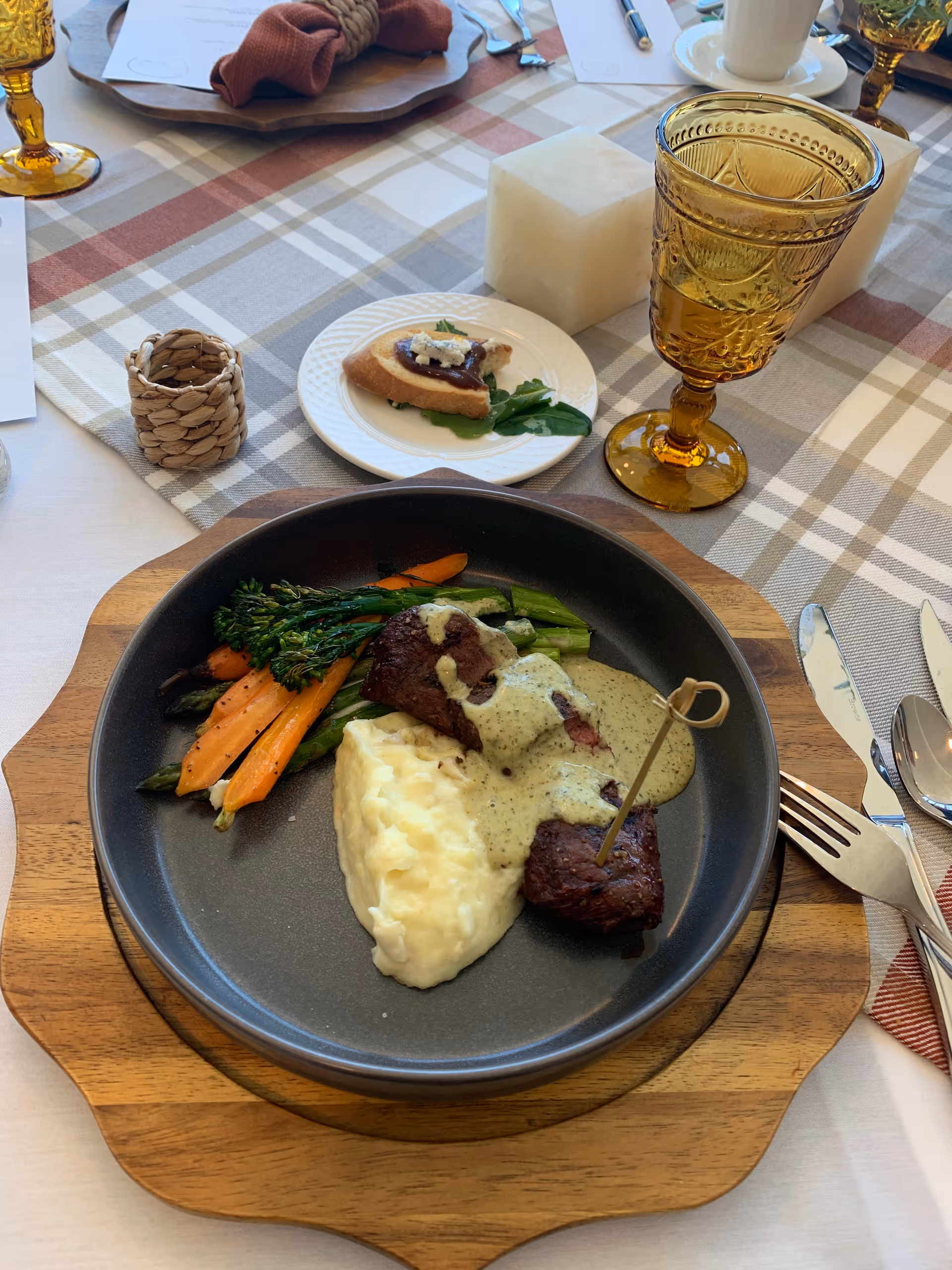 A plated meal on a wooden serving board featuring mashed potatoes, grilled vegetables including carrots and broccolini, and two pieces of steak topped with a creamy herb sauce. The table is set with a plaid tablecloth, amber-colored glassware, silverware, a small plate with a piece of bread and greens, and decorative candles.