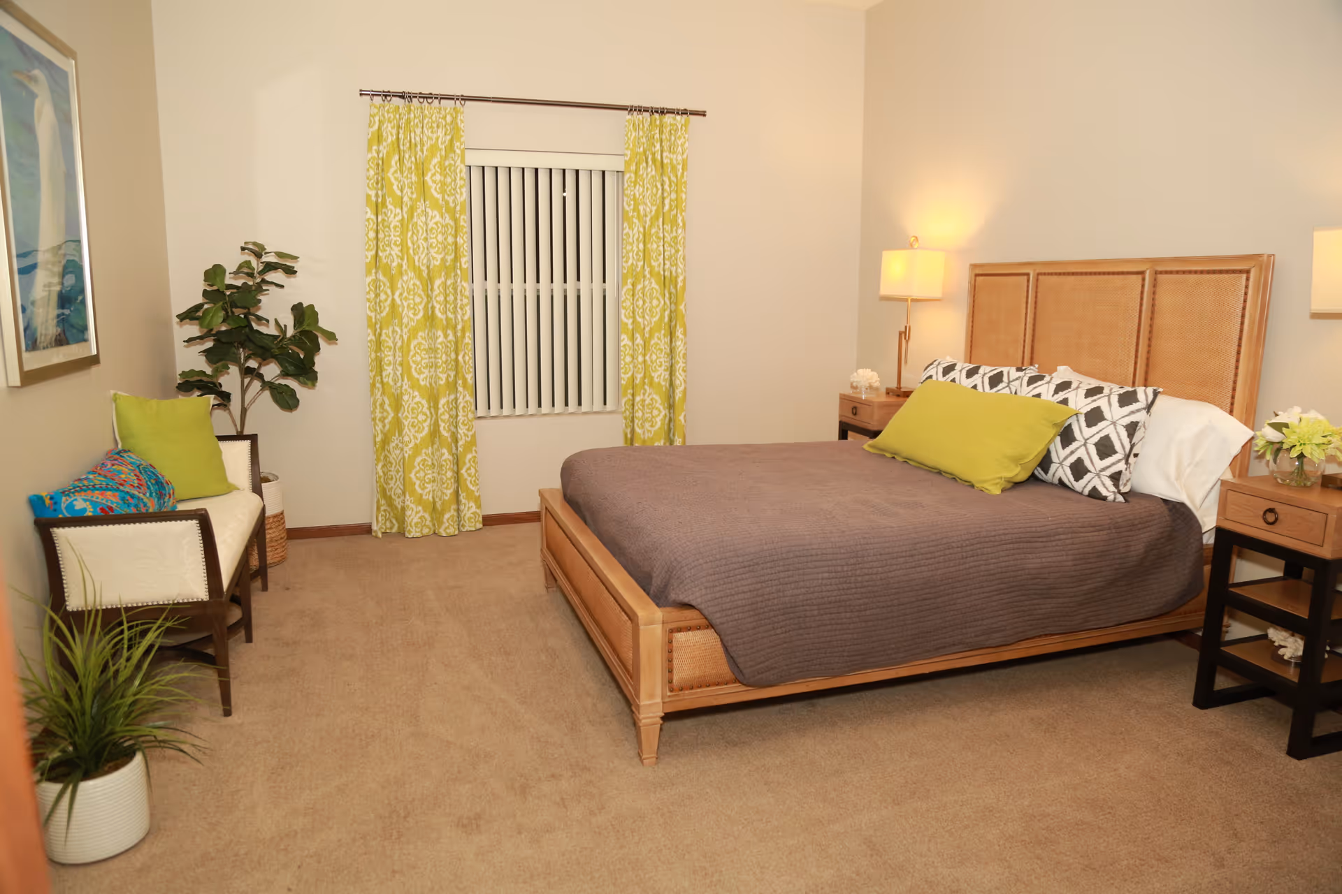 A cozy bedroom with a wooden bed frame and a brown bedspread, accented by green and patterned pillows. There are two wooden nightstands with lamps and decorative items on either side of the bed. A window with vertical blinds and yellow patterned curtains is centered on the far wall. To the left, there is a small bench with colorful cushions and a potted plant beside it. The room has beige carpet and neutral-colored walls.