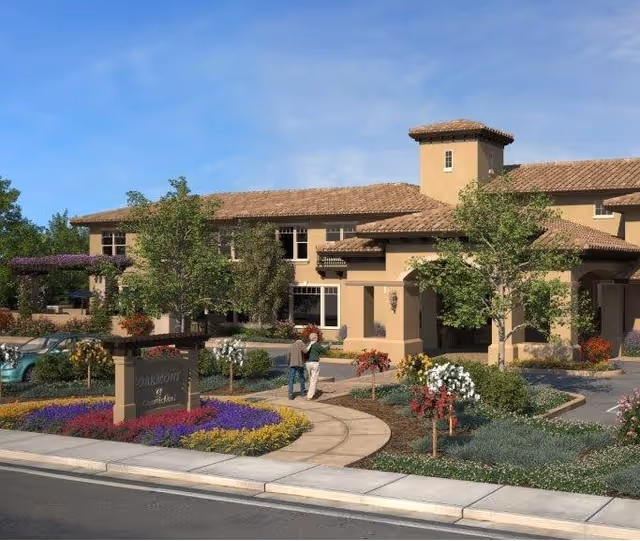 Exterior view of a senior living facility named Oakmont of Carmichael with a beige stucco building, tiled roof, landscaped garden with colorful flowers, trees, and a curved walkway. Two people are walking on the path near the entrance.