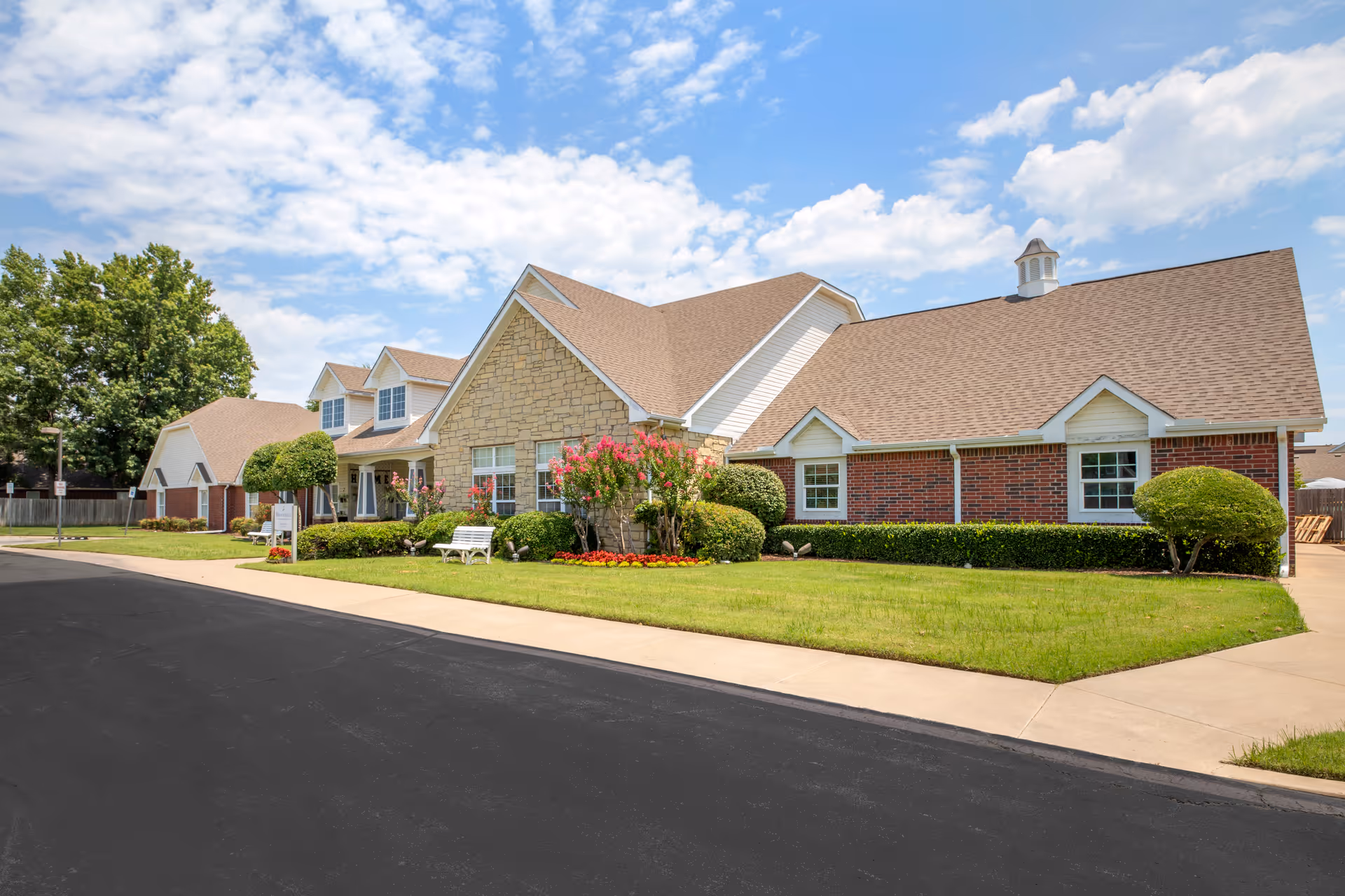 Exterior view of a senior living facility building with a combination of stone and brick walls, a well-maintained lawn, trimmed bushes, flowering plants, and a clear blue sky with some clouds.