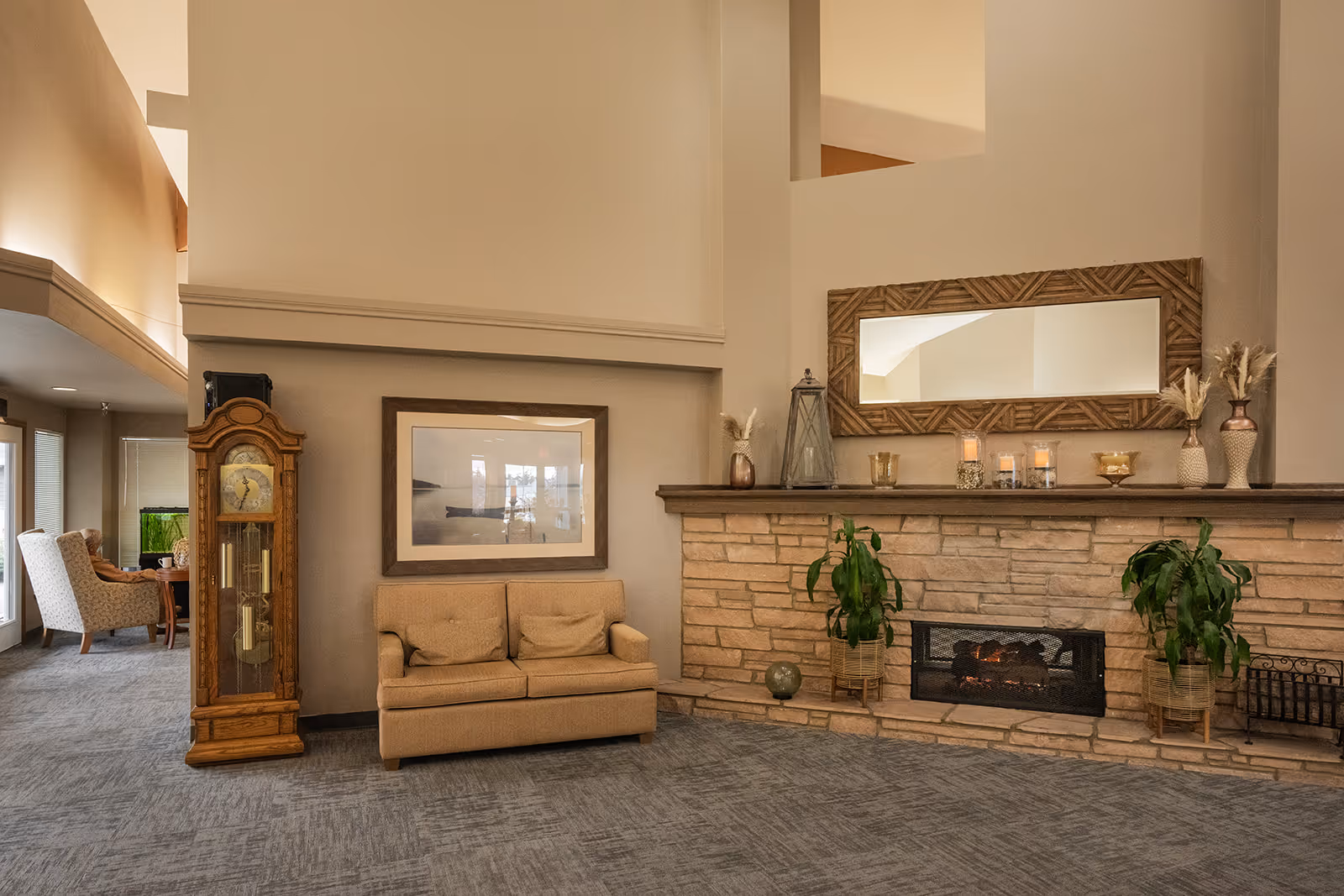 A cozy living room area featuring a stone fireplace with a wooden mantel decorated with candles, vases, and plants. Above the mantel is a large rectangular mirror with a wooden frame. To the left of the fireplace is a beige loveseat with two cushions, a framed picture on the wall, and a tall wooden grandfather clock. The room has carpeted flooring and neutral-colored walls, with additional seating visible in the background near large windows.