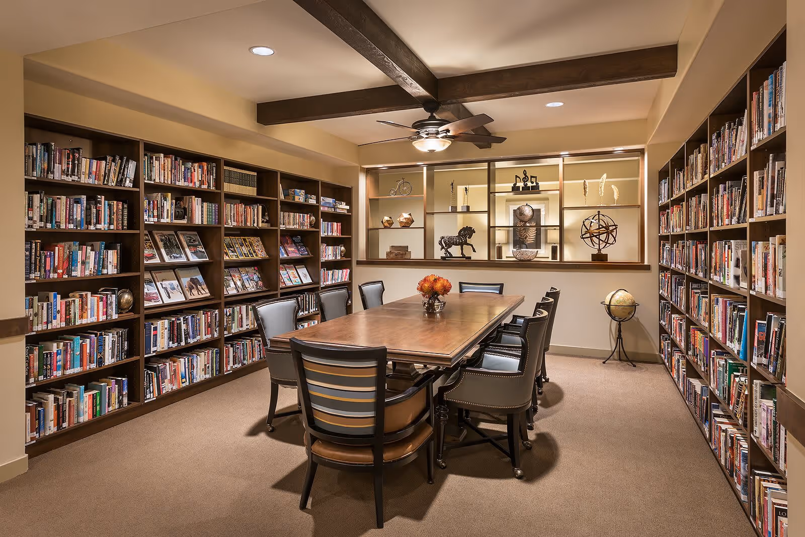 A cozy library room with wooden bookshelves filled with books lining both side walls. In the center, there is a large wooden table surrounded by eight chairs, with a small flower arrangement on the table. The ceiling has exposed wooden beams and a ceiling fan with a light. Decorative items are displayed on shelves on the far wall.