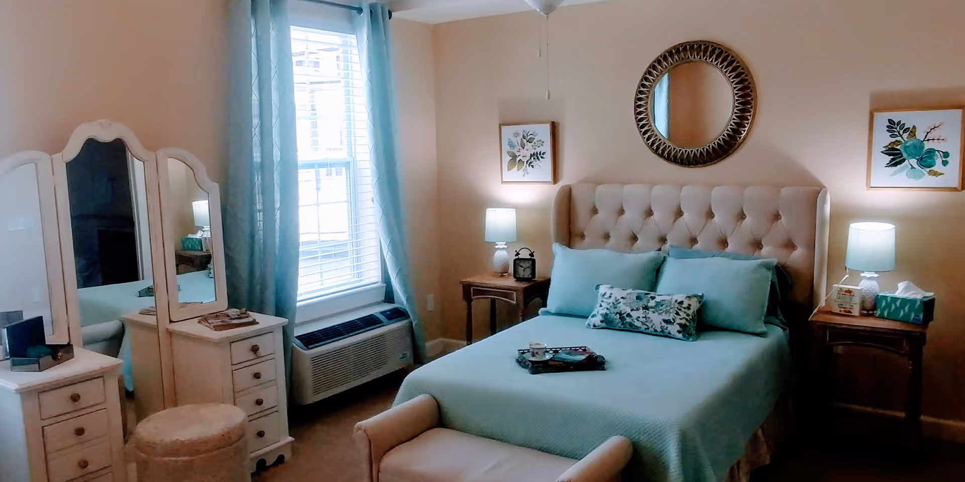 A cozy bedroom with a tufted headboard bed dressed in light blue bedding and decorative pillows. There are two wooden nightstands on either side of the bed, each with a lamp and decorative items. A round mirror hangs above the bed, flanked by two framed floral artworks. To the left, there is a white vanity with a three-panel mirror and a cushioned stool. Light blue curtains frame a window with blinds, and an air conditioning unit is installed below the window.