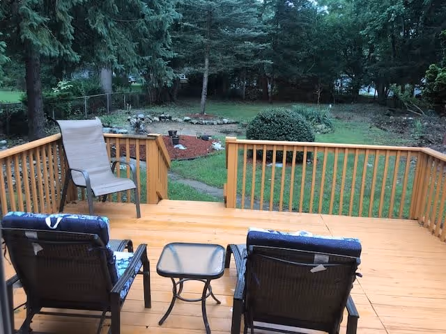 Wooden deck with two cushioned chairs and a small glass-top table, one additional chair without cushion, overlooking a grassy backyard with trees, shrubs, and a stone-bordered garden bed.