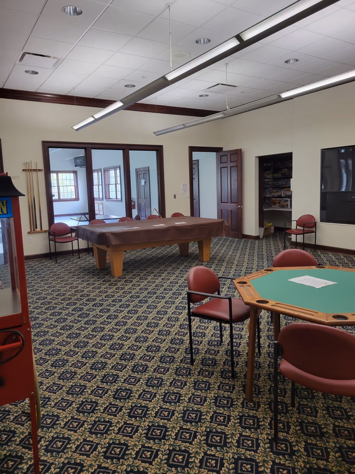 A recreational room with patterned carpet, a covered pool table, a green octagonal card table with four red chairs, and a wall-mounted television. There are windows and doors leading to adjacent rooms, one of which contains shelves filled with board games.