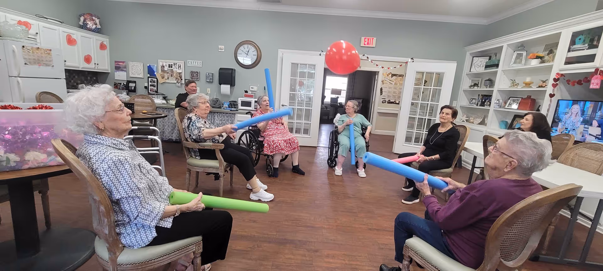 A group of elderly women seated in a circle in a common room, playing a game with pool noodles and a red balloon. The room has wooden floors, white cabinets, a clock on the wall, and a television. There are decorations with hearts, suggesting a festive or themed event.
