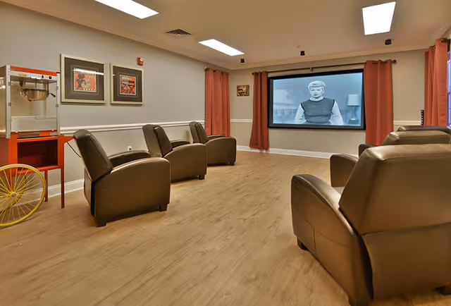 A cozy media room with six brown leather recliners arranged in two rows facing a large screen showing a black and white movie. The room has light wood flooring, beige walls with white trim, red curtains on the windows, and a popcorn machine on a red stand with yellow wheels in the corner.